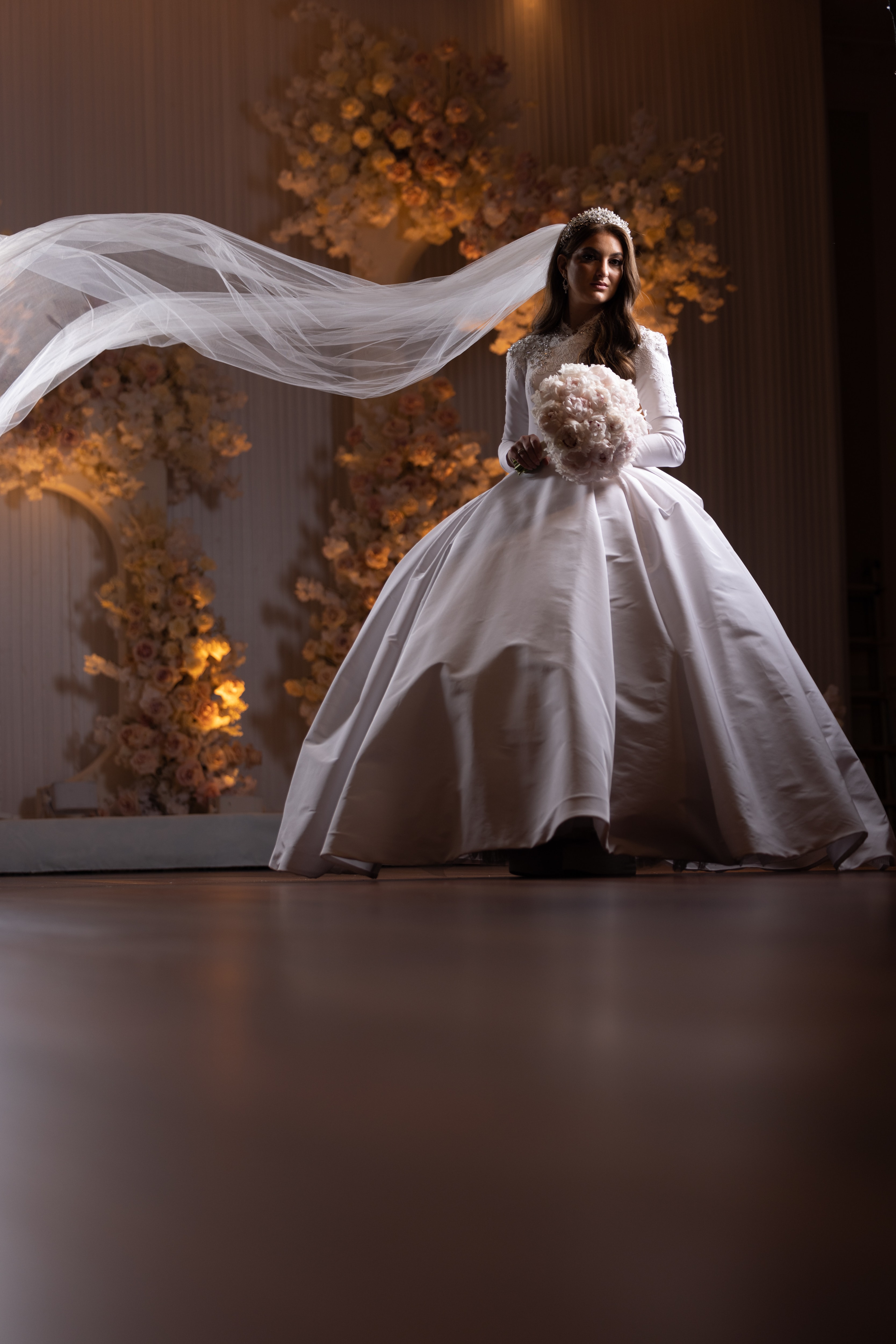Bride in satin gown with dramatic veil sweep against a warm-lit floral arch
